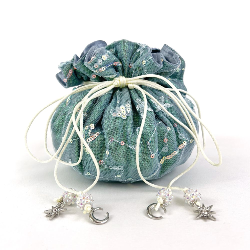 Exterior, side view shot of a closed dice bag. The bag is made of embroider, sequined netting over a shimmery blue, green material. The bag has white drawstrings with small moon and star charms on the ends and white sparkly beads. The bag stands upon a white backdrop.