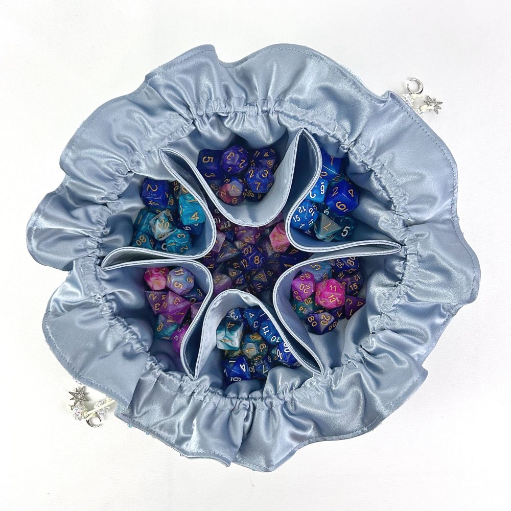 A top down view of an open dice bag. The interior of the bag is made entirely of a blue, grey satin. The bag has approximately 140 pink, blue and purple dice inside held within a large central section and six side pockets. The bag stands on a white surface. 