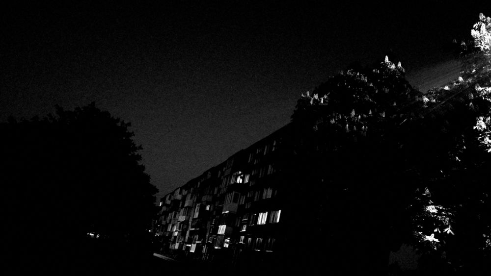 A monochrome photo of skewed perspective of a five story building and a couple of trees lit from the side by a street light.