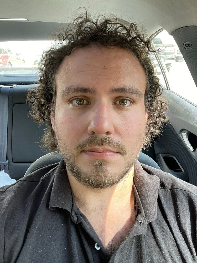 Incredibly tired and probably a little dehydrated in my black polo looking into the selfie camera while seated in the driver seat of my car. Without my glasses with my long hair. 