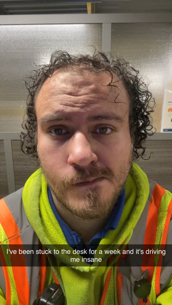 A selfie of me at work, in my slightly cold weather gear.  Looking absolutely pleased /s. The caption below my face says, "I've been stuck to the desk for a week and it's driving me insane"