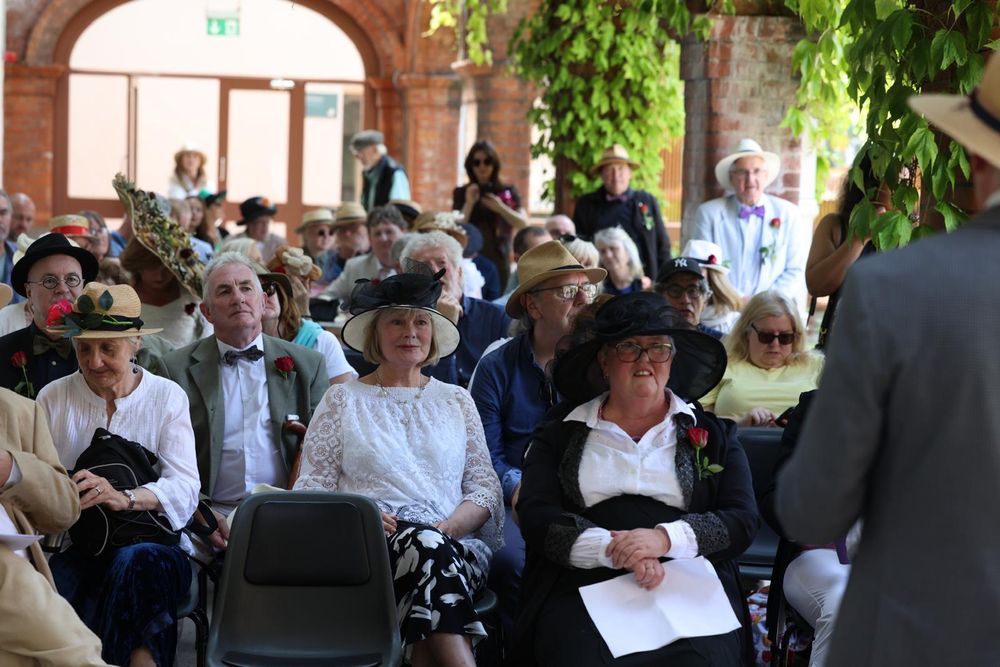 A seated crowd some of whom are dressed in large hats and suits with bowties
