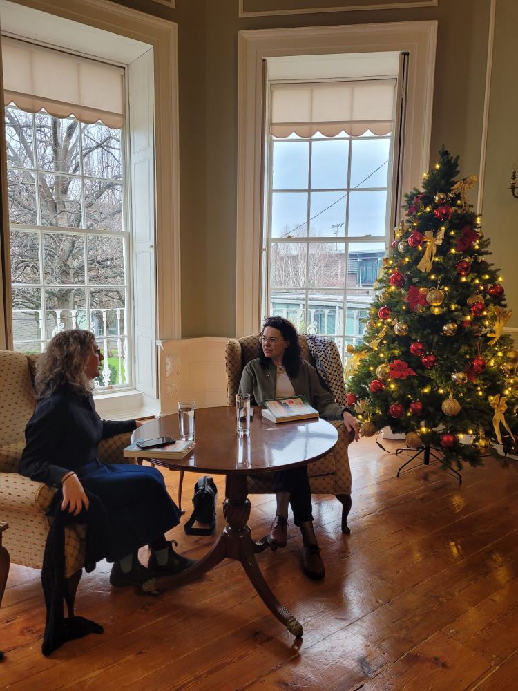 Juliana Adelman and Aingeala Flannery sit in wingback chairs on either side of a small round table in a bay window, with a Christmas tree to the right