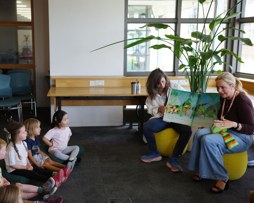 2 women sit on low yellow stools with a big colourful book between them They are reading it animatedly to a group of rapt children sitting in a semi circle