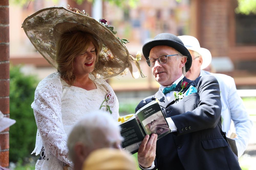 A woman with a large hat looks into a book held by a man in a suit and bowler hat