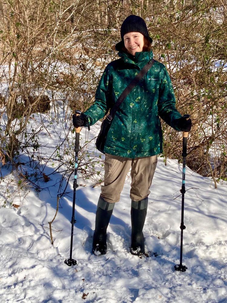 Woman (me) wearing a green ski jacket, tan pants and tall snow boots standing in snow and holding hiking poles