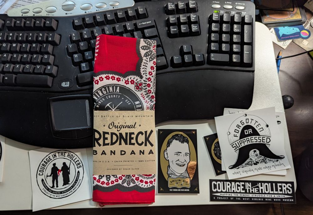 A collection of items is arranged on a desk in front of a black ergonomic keyboard. The centerpiece is a folded red bandana labeled "Original Redneck Bandana," commemorating the 1921 Battle of Blair Mountain in Mingo County, West Virginia. Around the bandana are several stickers and cards, including:

A circular sticker with two silhouetted miners and the words “Courage in the Hollers – Mapping the Miners' Struggle for a Union. A project of the West Virginia Mine Wars Museum.”

A stylized sticker of a tricorn hat with the words “Forgotten or Suppressed – West Virginia Mine Wars Museum.”

A rectangular sticker reading “COURAGE IN THE HOLLERS” in bold white letters on black.

A gold-framed illustration of a smiling man labeled “Smilin’ Sid – 1921 – West Virginia Mine Wars Museum.”


