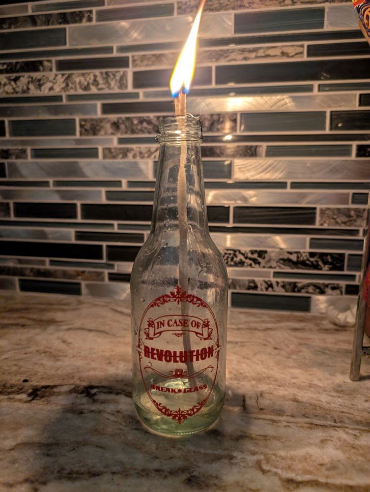 A glass bottle with a filigree design in red vinyl that reads In Case of Revolution Break Glass. It is being used as an oil lamp with a lit flame.