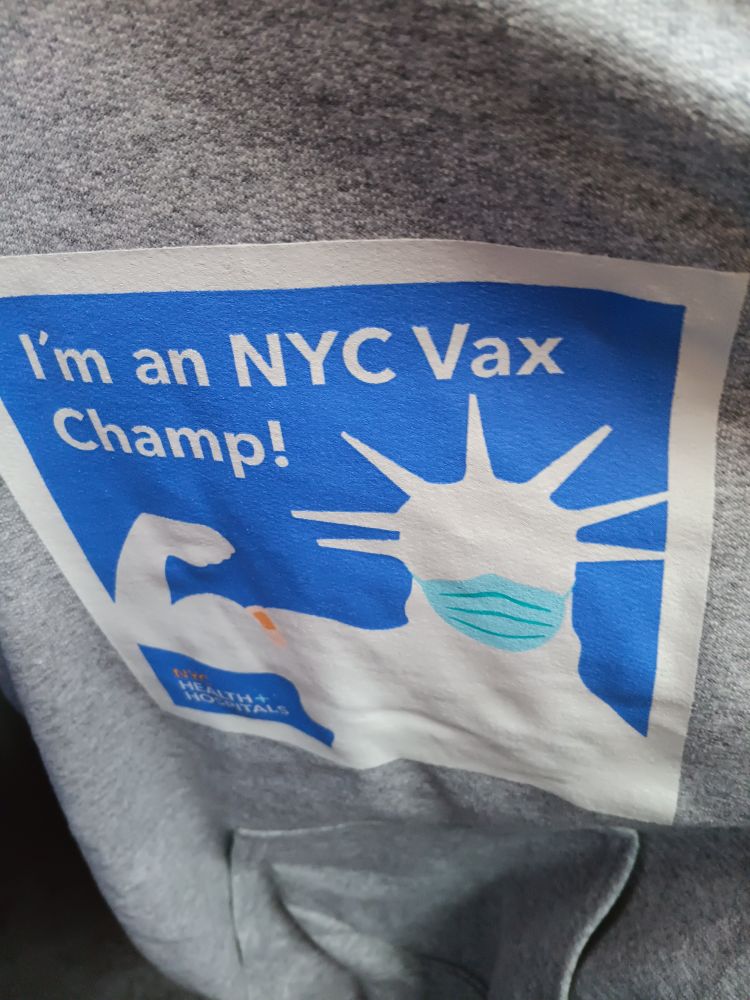 A blue and white graphic on a grey sweatshirt, showing a sihouette of the statue of liberty, masked, flexing an arm with a bandaid. Text says "In an NYC Vax Champ!"