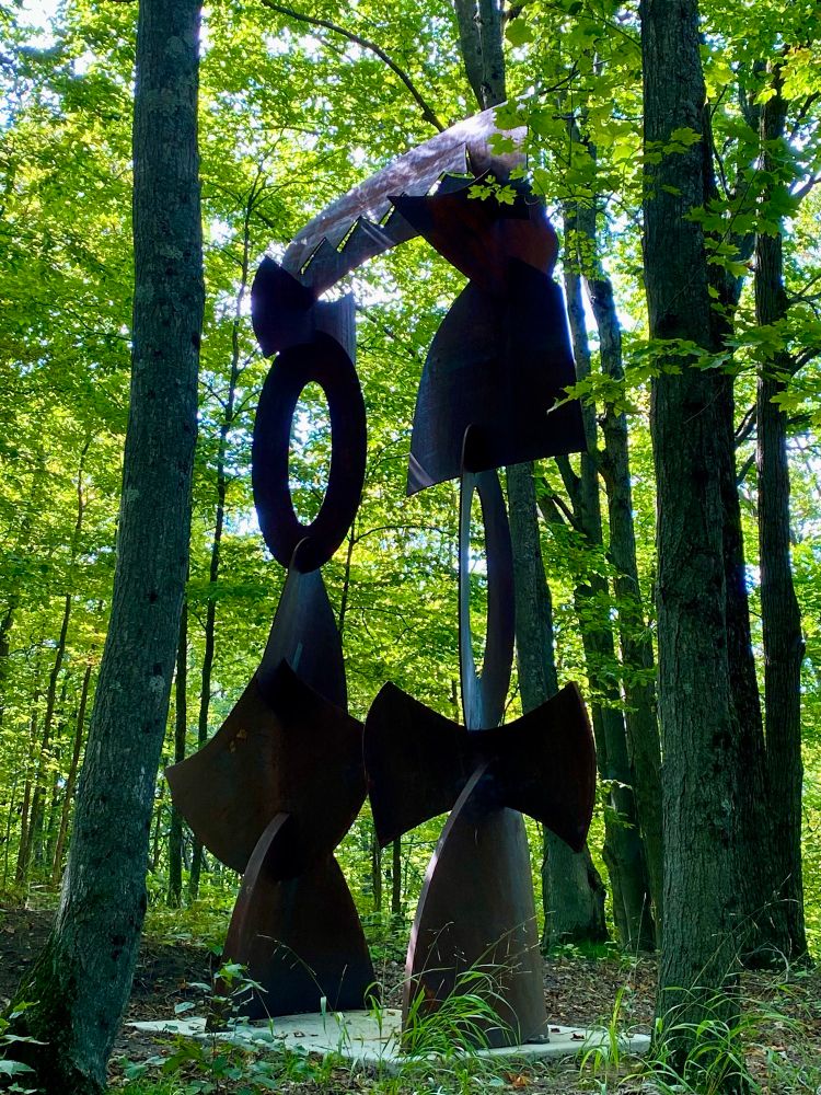 A large metal sculpture creating a gateway out of abstract shapes in the woods. 