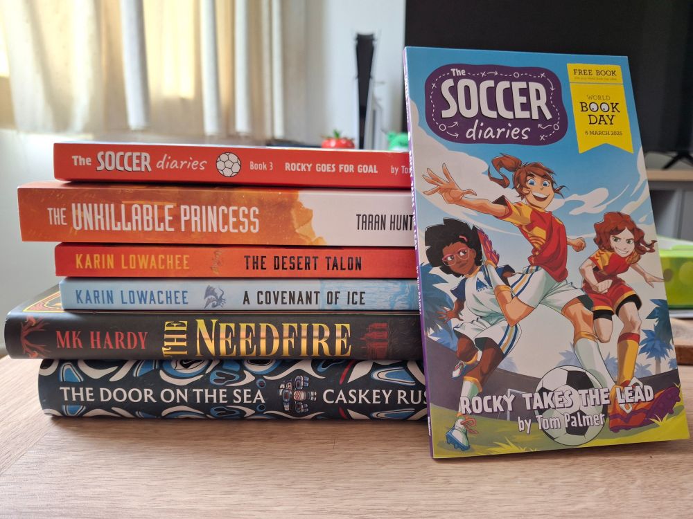 A stack of books on their side, top to bottom: The Soccer Diaries book 3, The Unkillable Princess, The Desert Talon, A Covenant of Ice, The Needfire, The Door on the Sea. The world book day Soccer Diaries Rocky Takes the Lead leans against the stack.