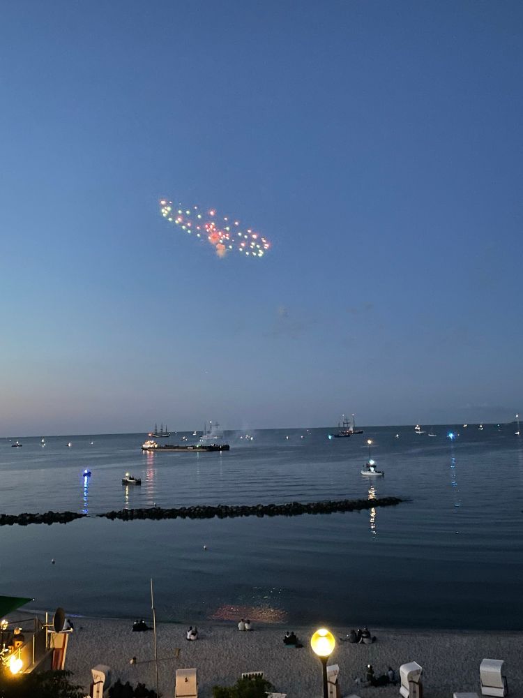 Viele Schiffe liegen vor Schilksee. Am Nachthimmel ein UFO-förmiger Rest eines Feuerwerkskörpers
