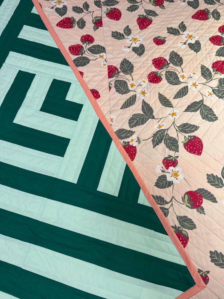 A detail photo showing the front and back of a handmade quilt. The front is a geometric design in two shades of green. The back is a strawberry print with green leaves, white and yellow flowers, on a light pink background. 