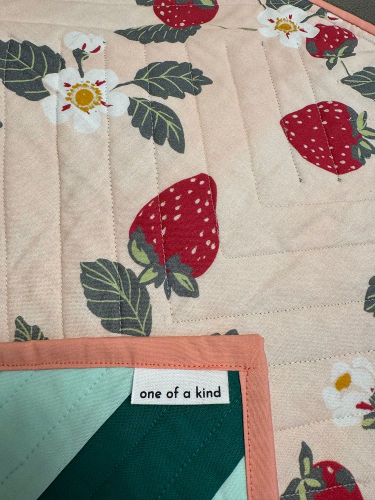 A detail photo showing the front corner and back of a handmade quilt. The front is a geometric design in two shades of green with a white tag that reads one of a kind. The back is a strawberry print with green leaves, white and yellow flowers, on a light pink background. 