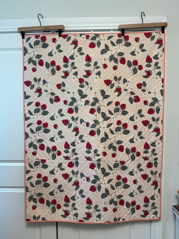 Photo of the back of a handmade quilt with a print of red strawberries, green leaves, and white flowers on a light pink background. 
