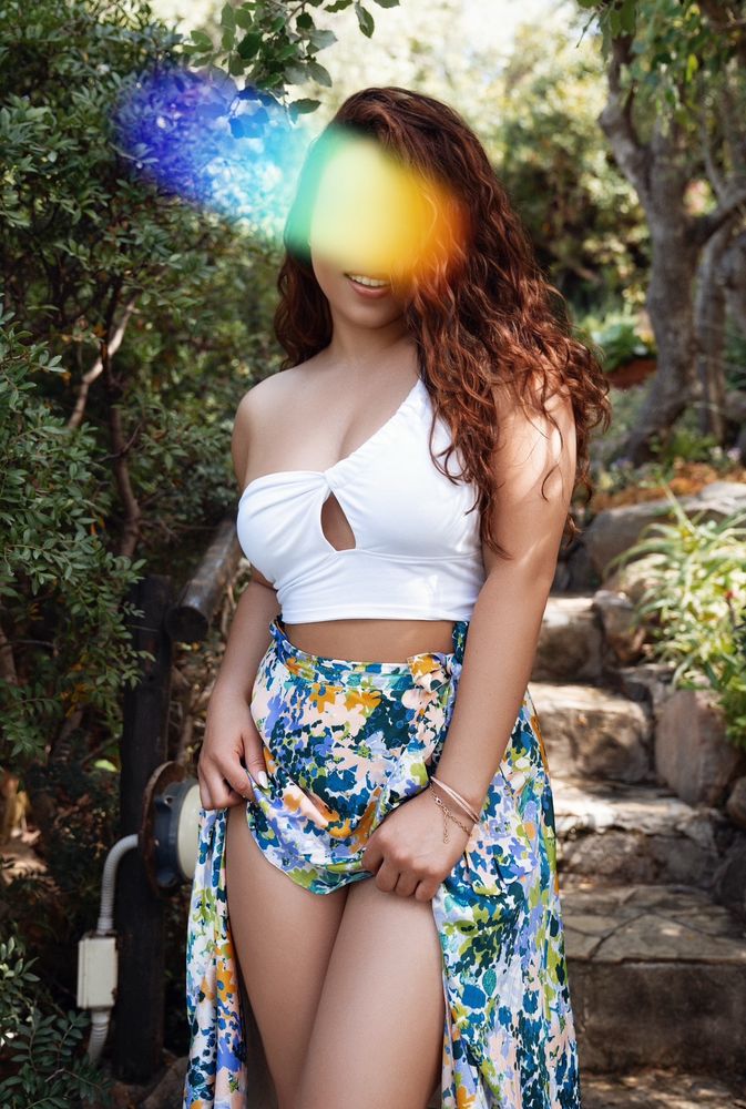 A smiling woman stands outside surrounded by greenery, lifting her skirt to show off her thighs.