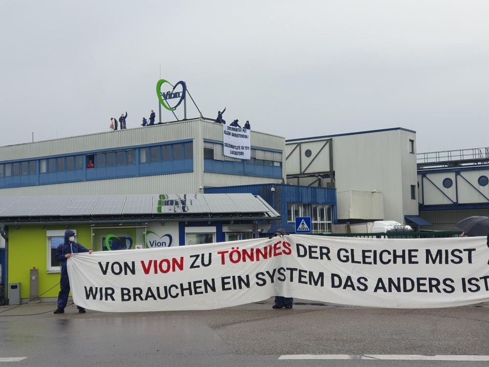 Protestaktion vor einem Vion-Schlachthof: Mehrere Personen blockieren den Eingang mit einem Banner auf dem steht "Von Vion zu Tönnies der gleiche Mist - Wir brauchen ein System das anders ist". Auch auf dem Dach protestieren Aktivist*innen mit einem Banner.