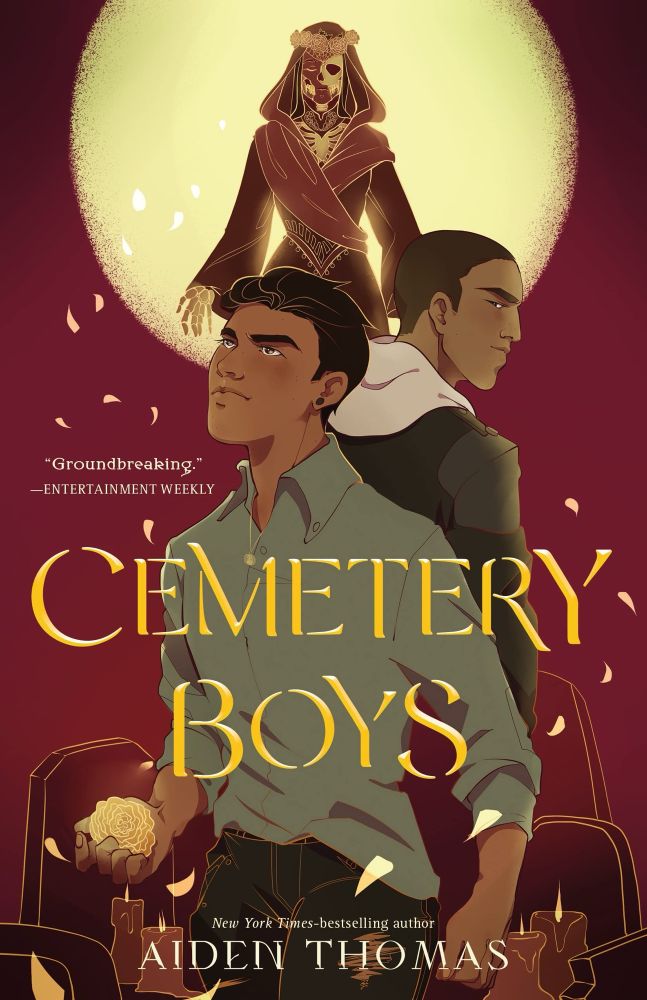 Two Latino boys stand back to back, one holds a glowing marigold, gravestones and candles are behind the lower half of them, and the moon covered partially by Death draped in a red cloak with a crown of marigolds on their golden, skeletal form is behind their heads. Over the boys is golden test that reads Cemetery Boys and below it white test reading New York Times bestselling author Aiden Thomas
