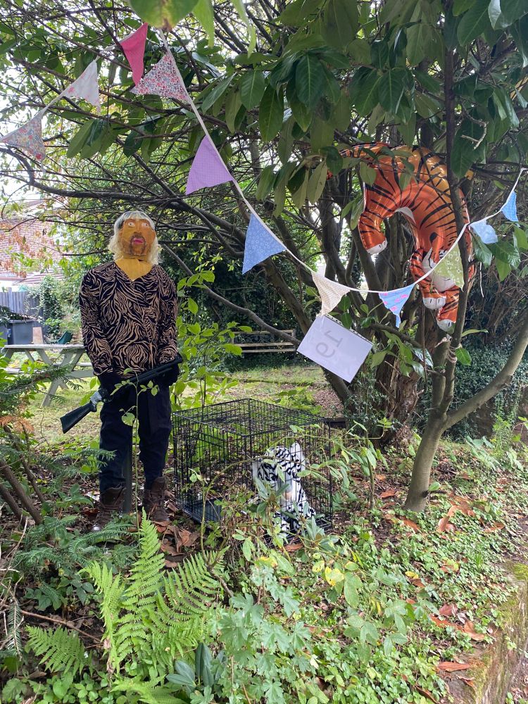A scarecrow of the Tiger King, including bright orange face paint, a leopard skin shirt, and bleach blond mullet. There is a stuffed tiger toy in a cage to his left, and a bright orange tiger balloon in a tree.