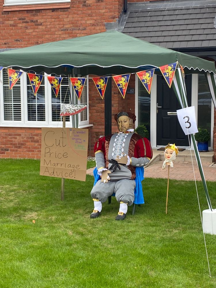 A scarecrow of King Henry VIII, dressed in ornate royal red. A sign to the left of him reading ‘Cut Price Marriage Advice!’ with an appraisal from ‘A. Boleyn’: ‘I wouldn’t use anyone else!’ On the right is a doll’s head with a crown on a spike.