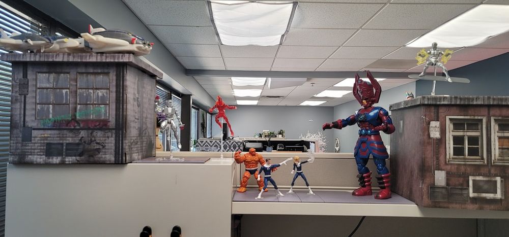 Marvel Legends on a cubicle shelf, with a cardboard building at the far end. A Silver Sufer figure is posed "flying" over the building, with a Galactus figure looming over Ben, Reed, and Sue, and Johnny "flying" over them at the far end. There is also a Rom action figure behind Johnny, another building, and the Fantasticar toy parked on top of that building.