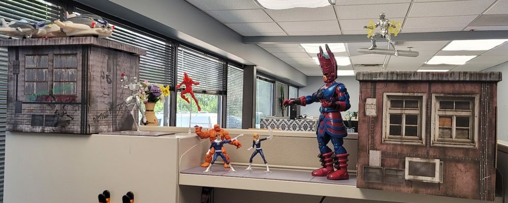 A wide shot of the cubicle shelf, with cardboard buildings on both ends. A Silver Surfer action figure is posed on top of one building, and a Fantasticar Toy is parked on the roof of another. Johnny and Rom action figures are posed in flight to the side of one building, and a 19" Galactus figure is posed in front of the other building. In between are Sue, Reed, and Ben, looking up at the Galactus figure.