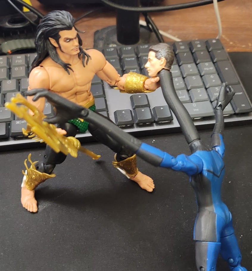 A 90s Namor Marvel Legends figure holding the extended neck of the 2005 Reed Richards Fantastic Four action figure in front of a keyboard on a desk.