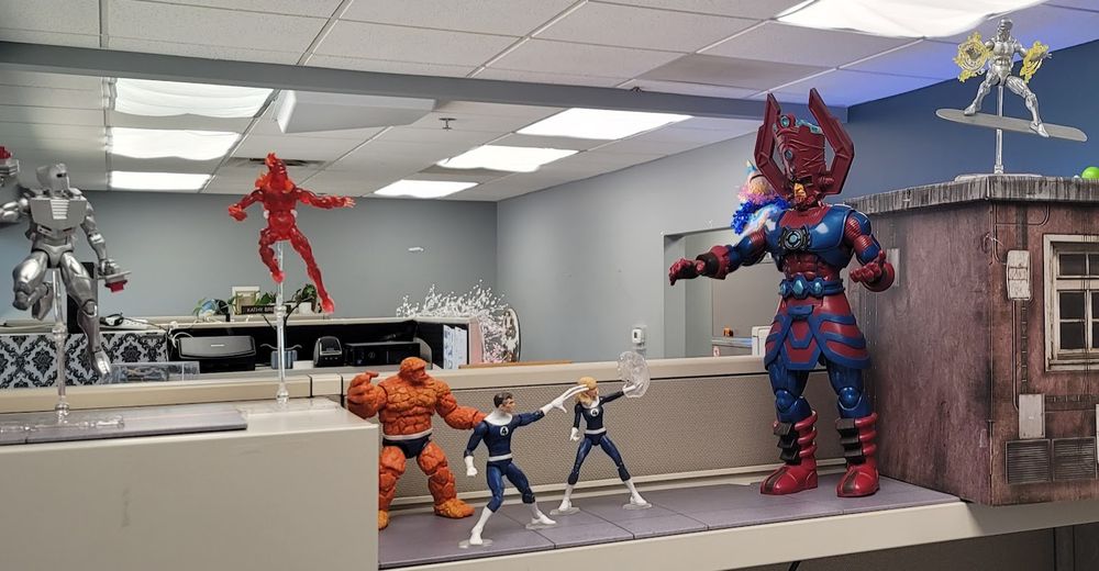 Marvel Legends on a cubicle shelf, with a cardboard building at the far end. A Silver Sufer figure is posed "flying" over the building, with a Galactus figure looming over Ben, Reed, and Sue, and Johnny "flying" over them at the far end. Behind Johnny, a Rom action figured is also posed in "flight."