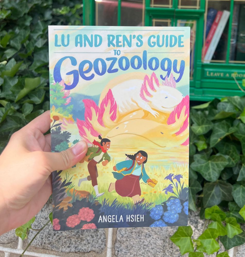 Photo of the author holding a copy of her middle grade graphic novel, LU AND REN'S GUIDE TO GEOZOOLOGY. The cover features two 12 year old girls running across a grassy lakeside with giant smiling axolotls in the background. 