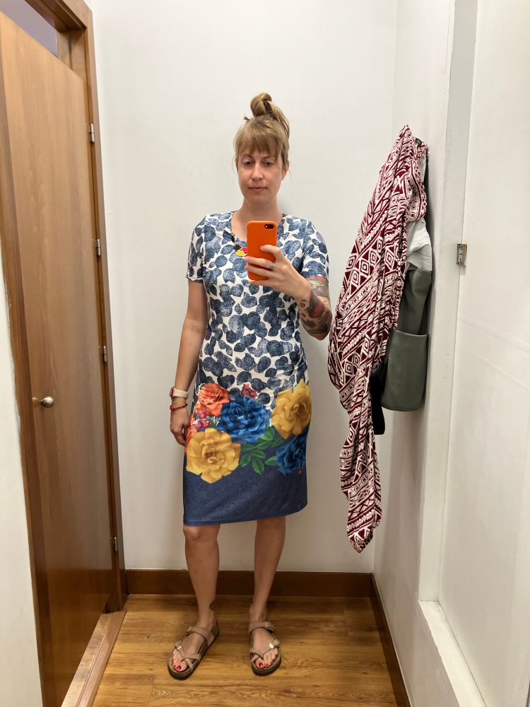 me in a dressing room, wearing a dress with shoulder pads, a sewn-on necklace detail, a denim-colored leaf pattern on the top half, and large, colorful flowers printed on the bottom half
