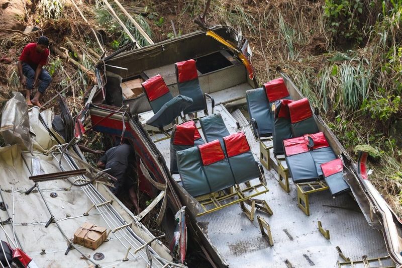 An aerial view of a bus crash site in Kotmale, Sri Lanka, shows a damaged vehicle on its side, surrounded by dense vegetation and uneven terrain. The bus's interior is partially visible, revealing several seats with red and blue covers, some of which are flipped or dislodged due to the impact. Two individuals can be seen working near the wreckage, one climbing on the side of the bus while the other is bent down, inspecting the area. The scene conveys a somber mood, highlighting the tragic aftermath of the accident, which claimed 21 lives. The surrounding environment features tall grass and scattered debris, emphasizing the urgency for improved road safety reforms in the country after this devastating incident.