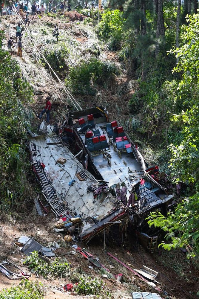 A devastated bus lies on its side in a steep, overgrown area, illustrating the aftermath of a tragic road accident. The bus is largely mangled, with several seats visible, some red and grey, scattered inside. Surrounding the vehicle are remnants of personal belongings, such as bags and items likely belonging to passengers. A group of onlookers and emergency responders can be seen in the background, some climbing the hillside, indicating a search and rescue effort following the accident. The scene is engulfed in dense greenery, with trees and tall grasses surrounding the area, emphasizing the isolated and rugged environment where the tragedy occurred. This tragic incident, referred to as the Kotmale bus tragedy, resulted in the loss of 21 lives, underscoring the urgent need for road safety reforms in Sri Lanka, where a person dies every three hours in road-related accidents.