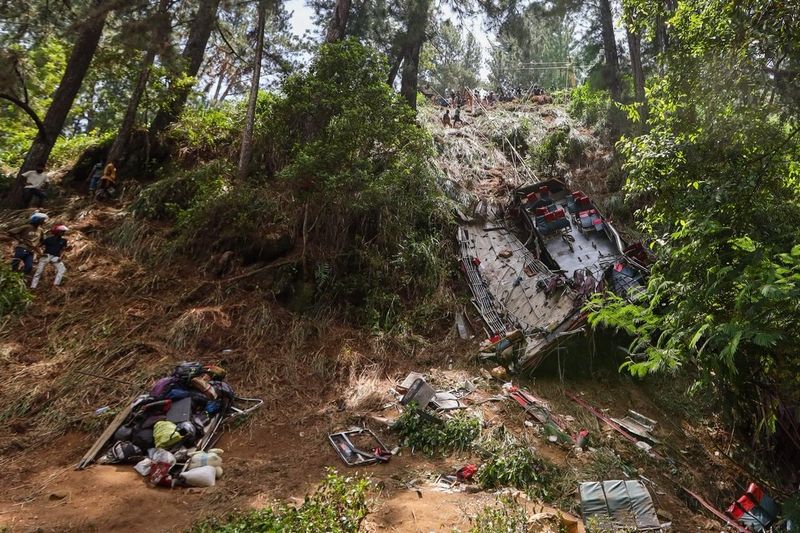 A devastating scene unfolds at the site of the Kotmale bus tragedy, where a bus has plunged down a steep, grassy embankment surrounded by tall trees. The bus is partially overturned, with its rear end visible, revealing empty seats and scattered belongings. The area is strewn with personal items, such as clothing and bags, indicating the chaos following the accident. Rescue workers and onlookers are seen navigating the slope, as they search for victims. The lush greenery contrasts sharply with the somber nature of the scene, emphasizing the urgency for road safety reforms in Sri Lanka, where road accidents claim a life every three hours. The image captures the stark reality of the tragedy, highlighting both the natural environment and the human impact of such events.