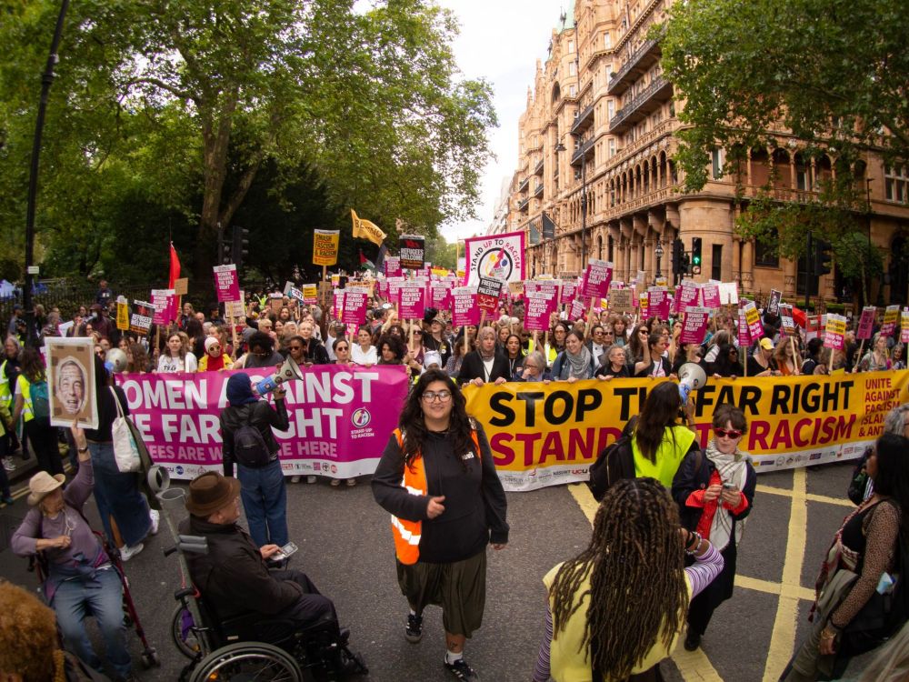 Wide shot of a massive, crowded anti-far right/anti-racism demonstration marching down an ornate city street. Protesters hold signs and a large pink and yellow banner reading, in part, "WOMEN AGAINST THE FAR RIGHT", "STOP THE FAR RIGHT STAND UP TO RACISM."