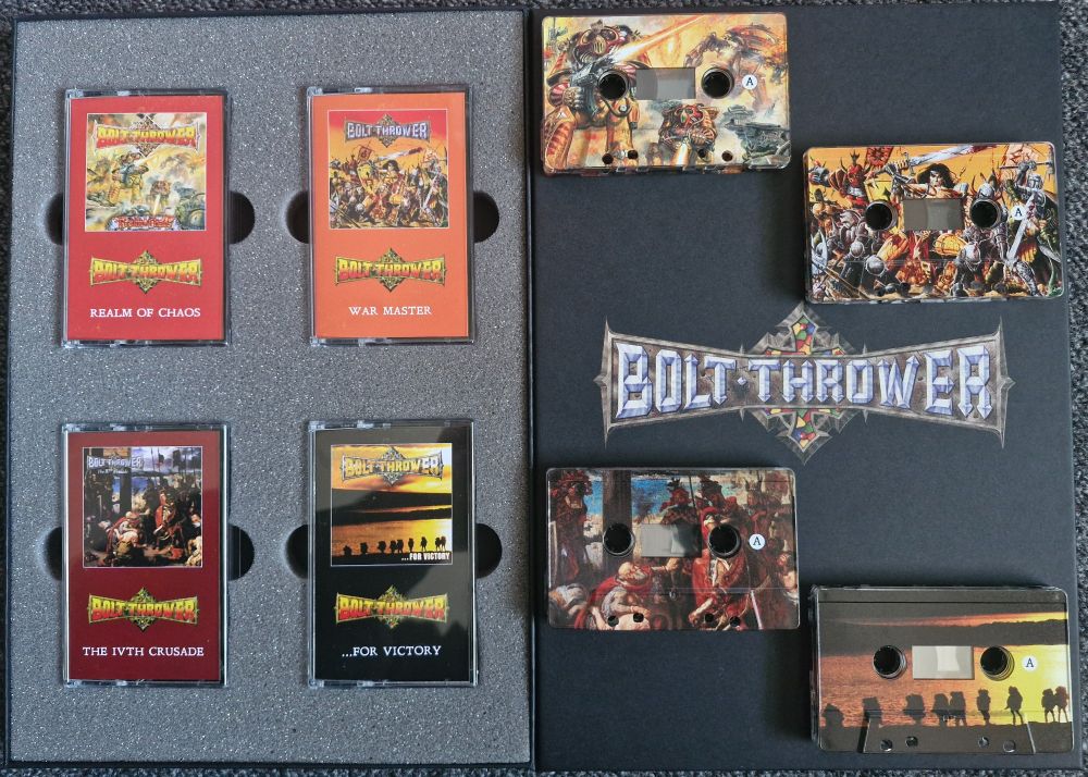 On the left side: 4 tape cases in a foam holder. On the right side: the 4 cassette tapes laid out on the box lid, with the Bolt Thrower logo on it.