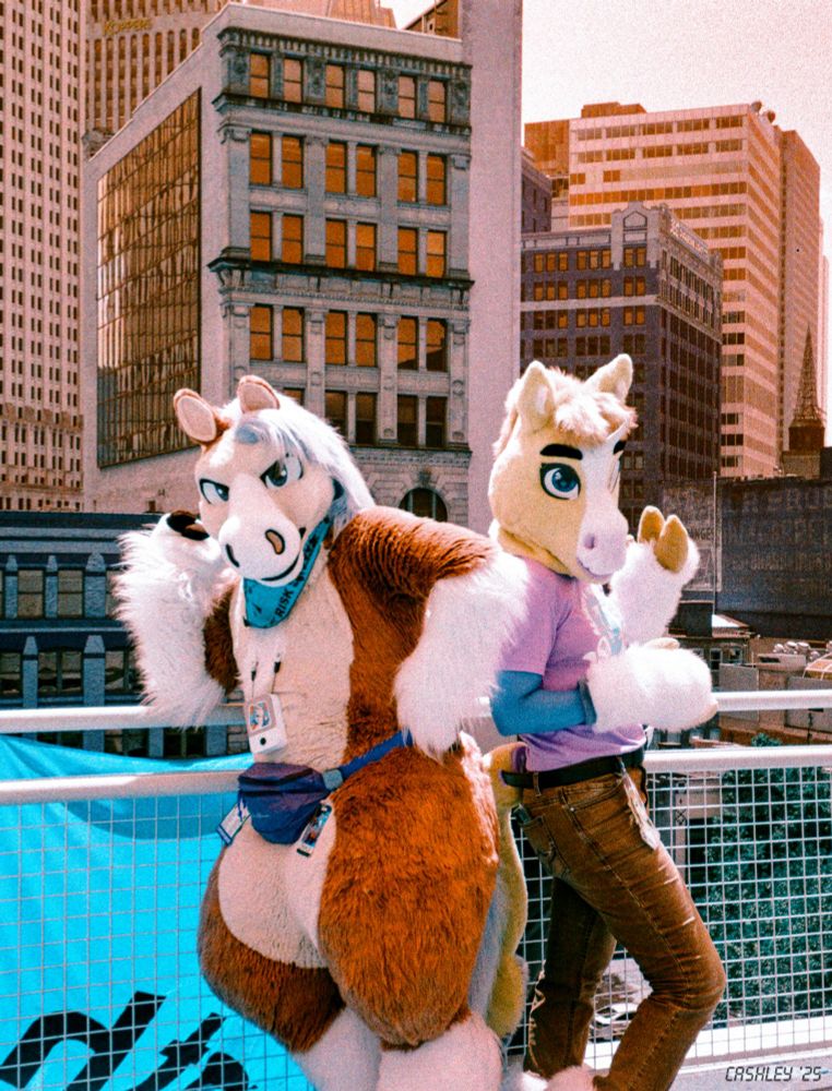 This is a photograph of a pegasus fursuiter and a kelpiecorn fursuiter taken using color-shifting film, so that what would normally be shades of blue and green are shifted to oranges and yellows and vice versa. They're standing outside on the rooftop of the DLCC with the Pittsburgh skyline in the background behind them, in a slightly dramatic pose that's almost but not quite the Charlie's Angels pose.