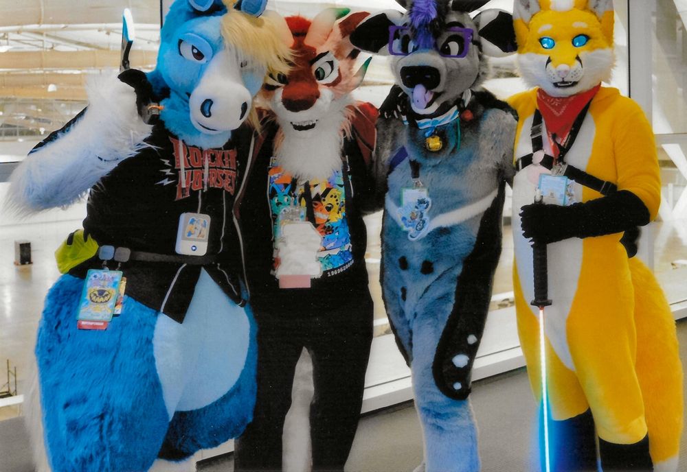 This is a film photograph of four fursuiters at Anthrocon 2024. From left to right is a blue and white pegasus, a red and pink dragon, a gray and purple deer, and a yellow and pink fox. 