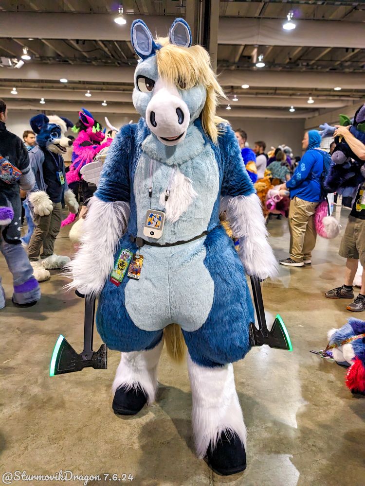 This is a photograph of a blue pegasus fursuiter standing in the cooldown hall following the 2024 Anthrocon fursuit parade, with other attendees and fursuiters in the background. He's holding a prop sci-fi tomahawk in each hand and directly facing the camera with his left hoof slightly forward.