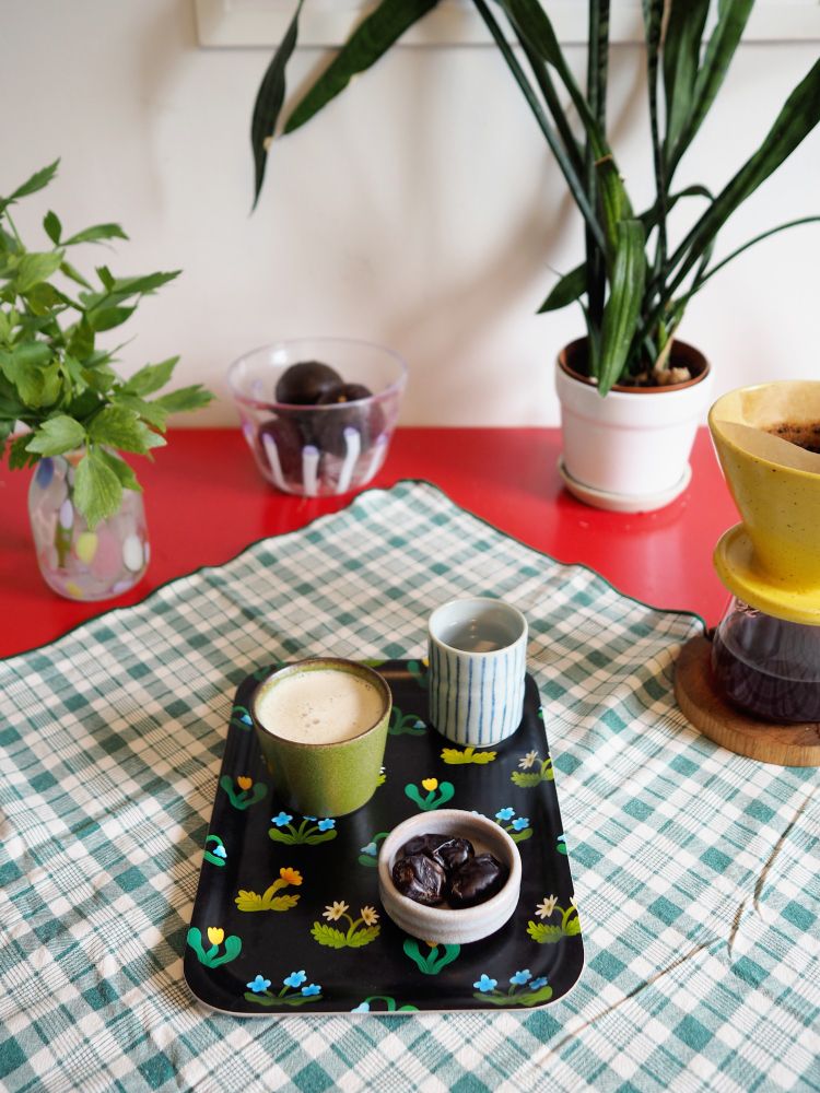 Another photograph of a small tray on a table. The tray has a floral pattern on a black background. On the tray are dates on a small dish and two cups, one with coffee and the other with water.