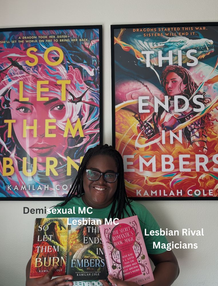 me holding up my books i have copies of in front of my poster size book covers with the words demisexual mc, lesbian mc, lesbian rival magicians near the books