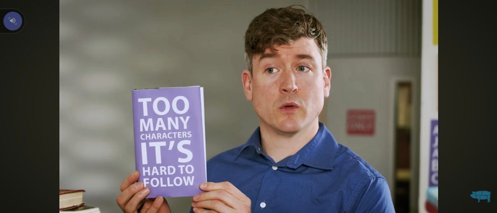 Screenshot from a video with a man holding up a book with a purple cover and white title: Too Many Characters It's Hard To Follow