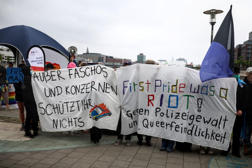 Menschen mit zwei Transpis stehen vor dem Polizeistand auf dem Straßenfest des CSD. Auf dem einen Transpi steht "Außer Faschos und Konzernen schützt ihr nichts". Auf dem Transpi ist ein brennendes Polizeiauto gemalt. Auf dem anderen Transpi steht "First Pride was a Riot! Gegen Polizeigewalt und Queerfeindlichkeit!"