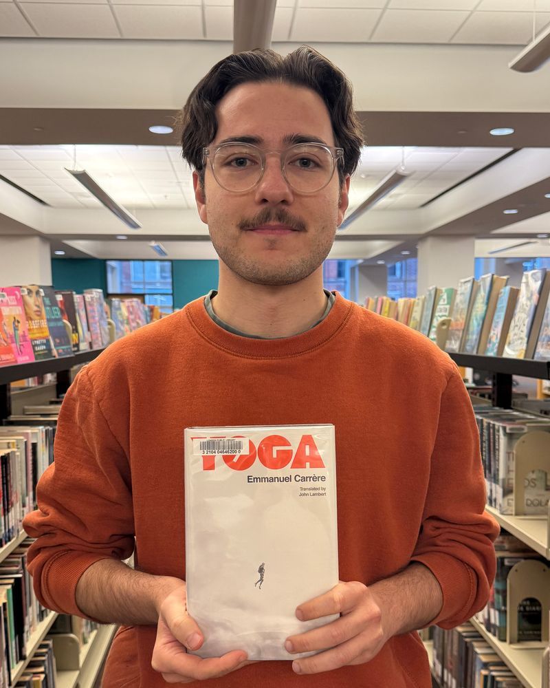 A staff member with a moustache and glasses wearing an orange sweater holding the book Yoga by Emmanuel Carrère.