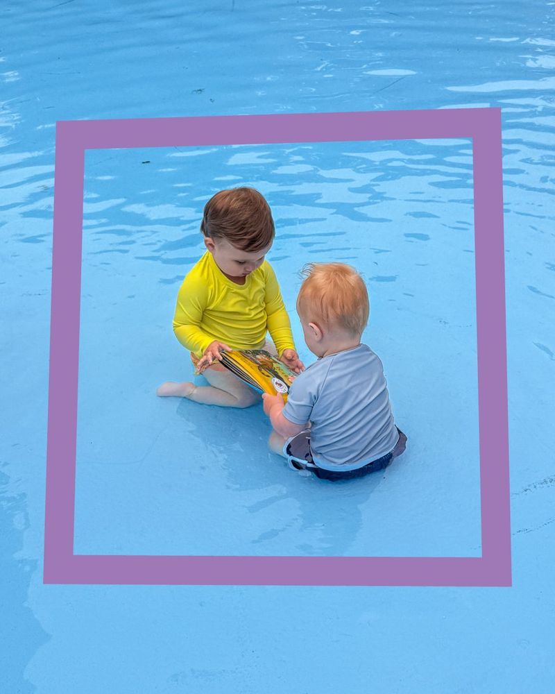 Two young children sit in shallow water, facing each other and looking at a book together. A purple rectangular border frames them.