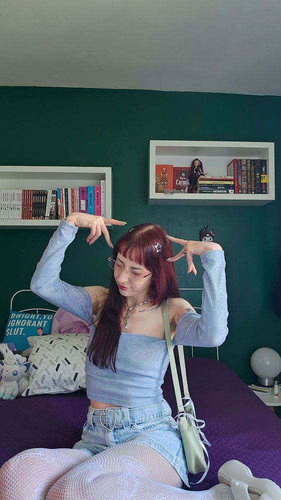 foto minha, mulher branca com cabelo vermelho posando, uso camiseta e shorts azuis, meia calça de renda e estou sentada em cima de uma cama.