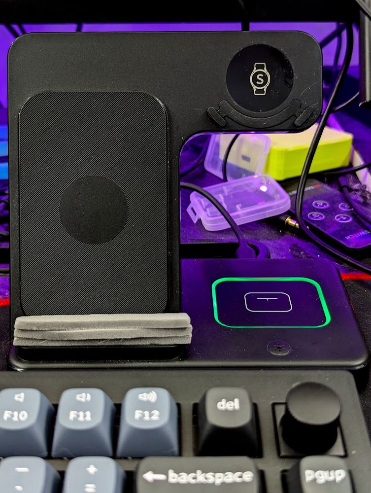 A wireless charging stand for phone and watch with a flat base, and an upright phone holder. The stand is black plastic. Three pieces of crudely cut foam with adhesive backing are stuck to the base where the phone would sit, in order to raise the height of the phone.