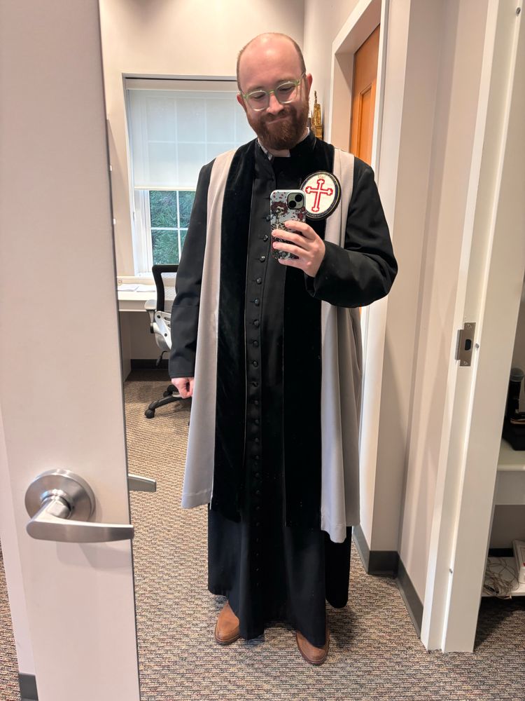 A person in the vestments of a verger and green glasses taking a mirror selfie