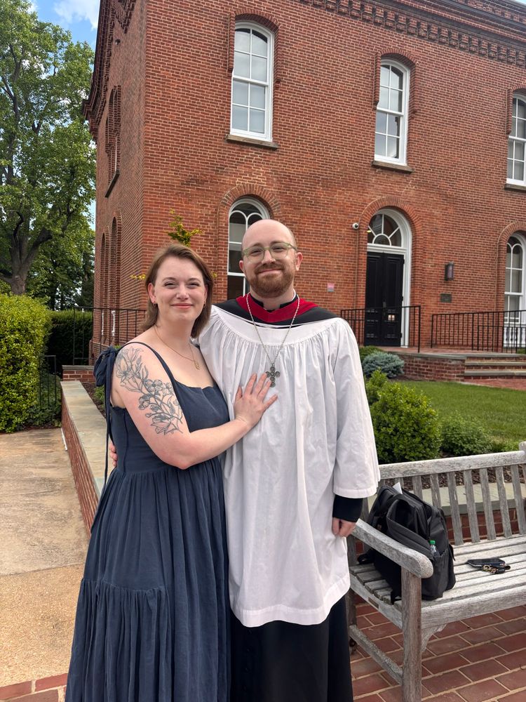 Two people side hugging. One in a linen dress and another in a cassock and surplice with necklace 