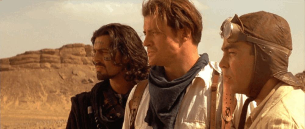 three men are standing next to each other in the desert and one of them is wearing a helmet