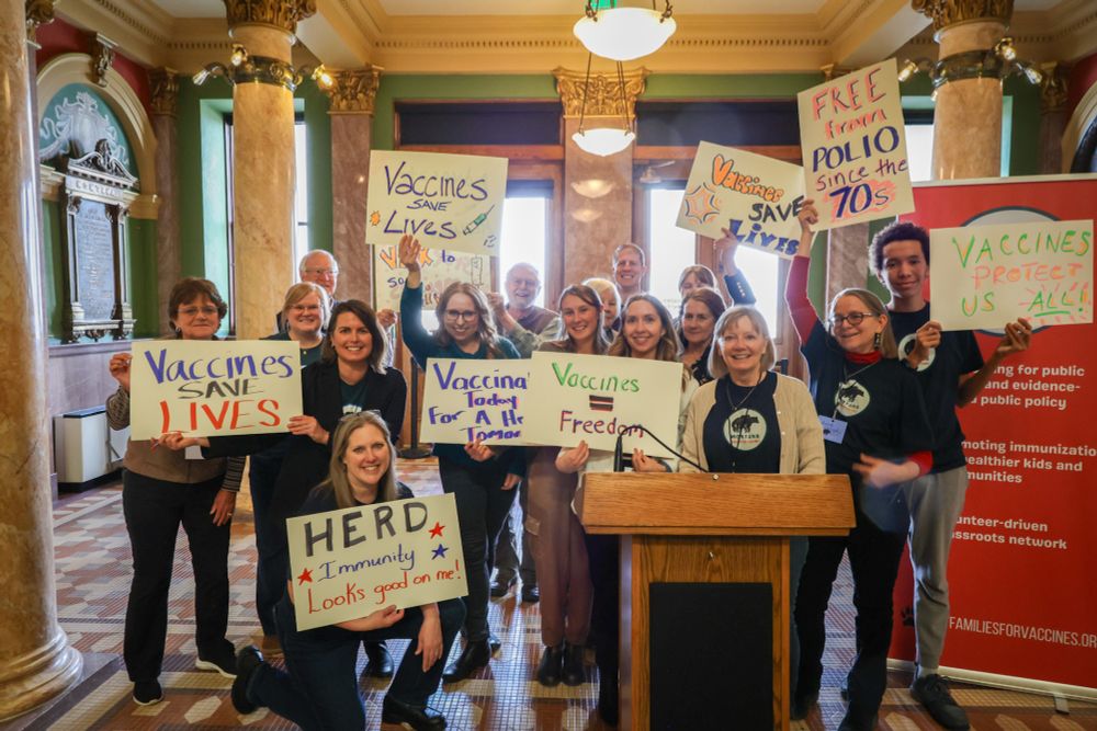 A group of approximately 20 people standing in the rotuda area of the Montana Capitol holding signs promoting vaccines. "Vaccines Save Lives" "Herd Immunity Looks Good on Me" and "Free From Polio Since the 70s"